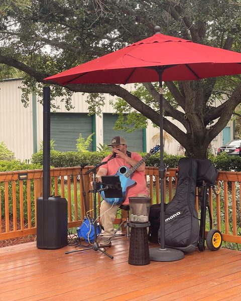 A musician playing guitar under a red umbrella on the deck