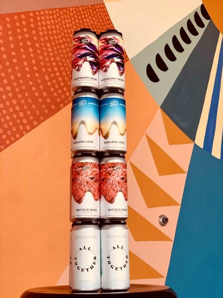 A stack of eight Brighter Days cans in front of a painted mural: Raspberry Beret, Endless Love, Bicycle Race, and All Together collaboration cans