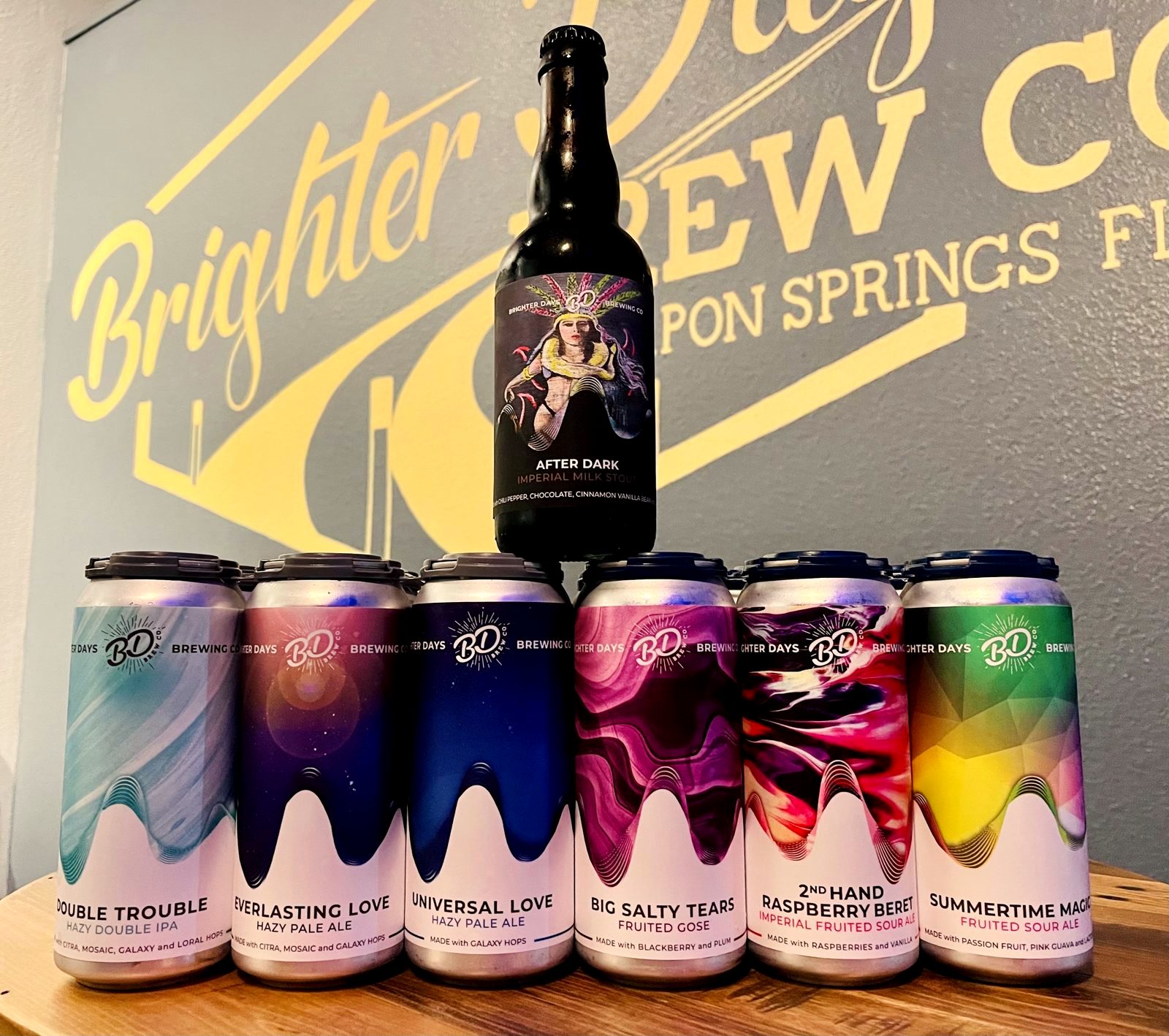 An After Dark bottle on top of six Brighter Days cans in front of the gold Brighter Days Brew Co. wall sign: Double Trouble, Everlasting Love, Universal Love, Big Salty Tears, 2nd Hand Raspberry Beret and Summertime Magic