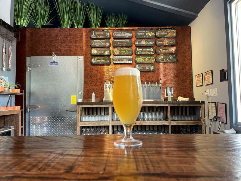 Glass of golden beer on a wooden table in the taproom, with menu signs on the brick wall behind