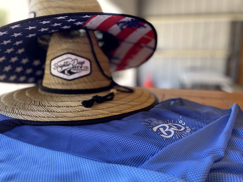 Straw hat with a Brighter Days patch and an American-flag brim, on a blue Brighter Days shirt