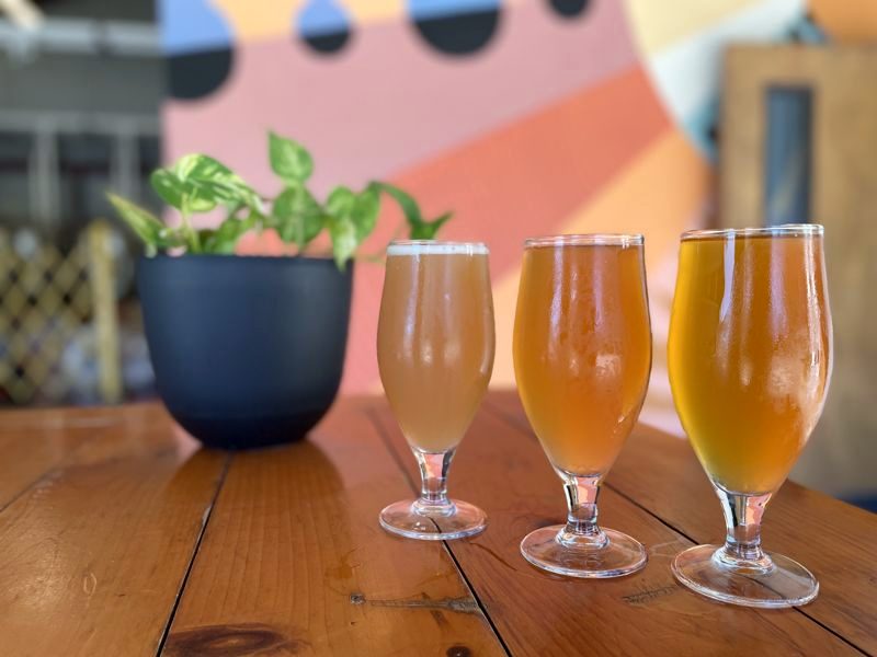 Three glasses of golden beer on a wooden table in front of a colorful painted mural