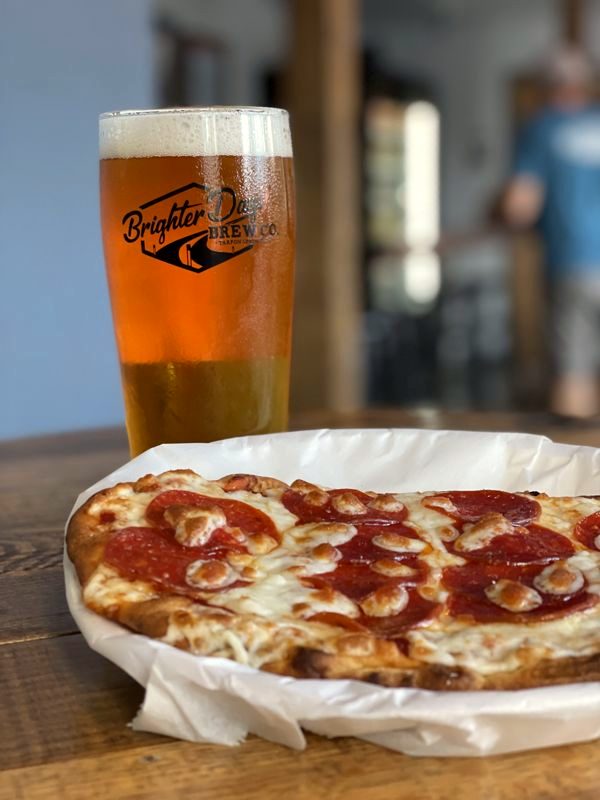 Pint of amber beer in a Brighter Days glass beside a pepperoni pizza on a wooden table