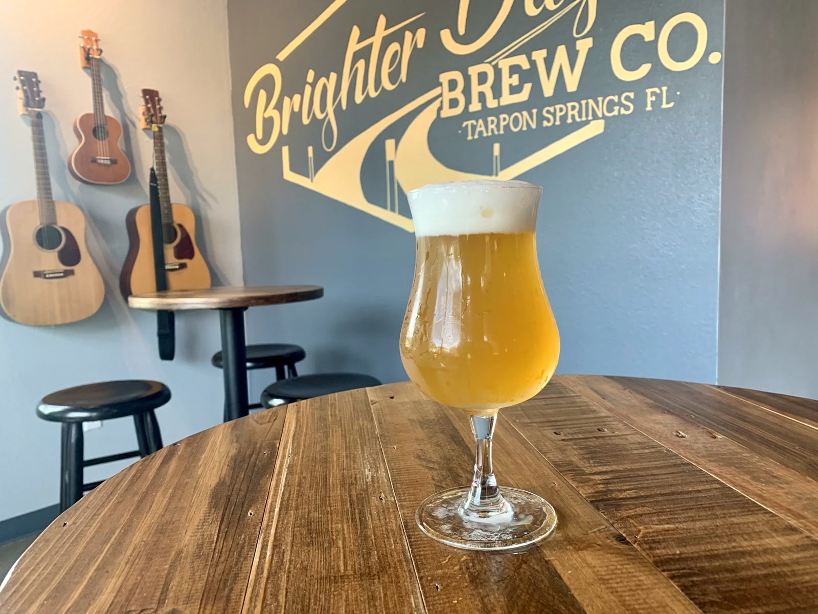 A tulip glass of hazy golden beer on a wooden table in front of the gold Brighter Days Brew Co. wall sign, with guitars and a ukulele hanging on the wall