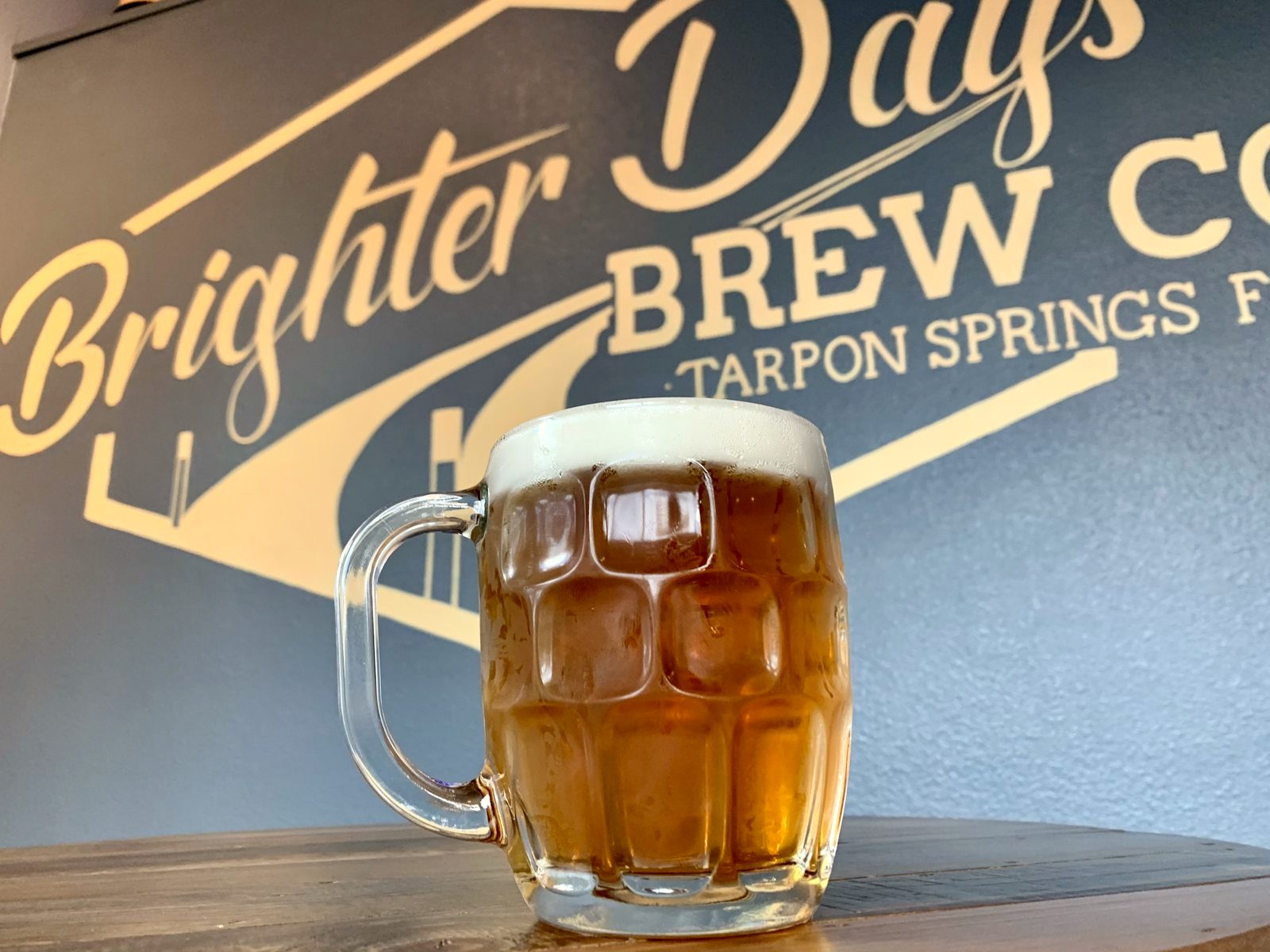 A glass mug of amber beer on a wooden table in front of the gold Brighter Days Brew Co. Tarpon Springs wall sign