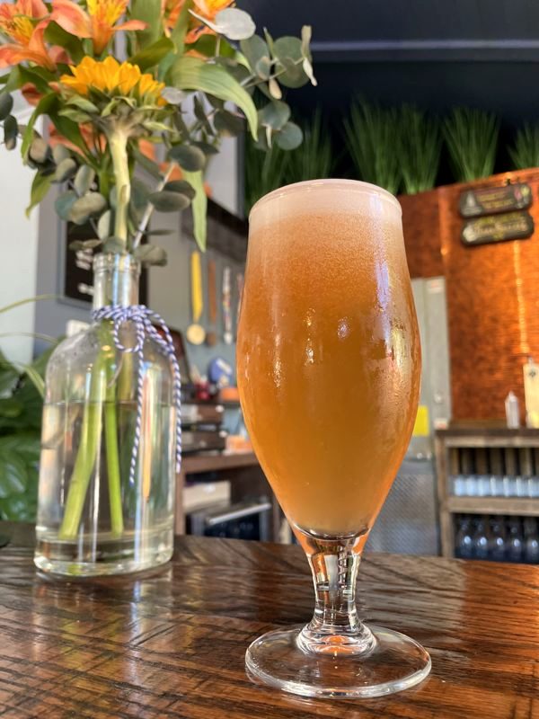 Tulip glass of hazy golden beer on a wooden bar next to a vase of orange flowers